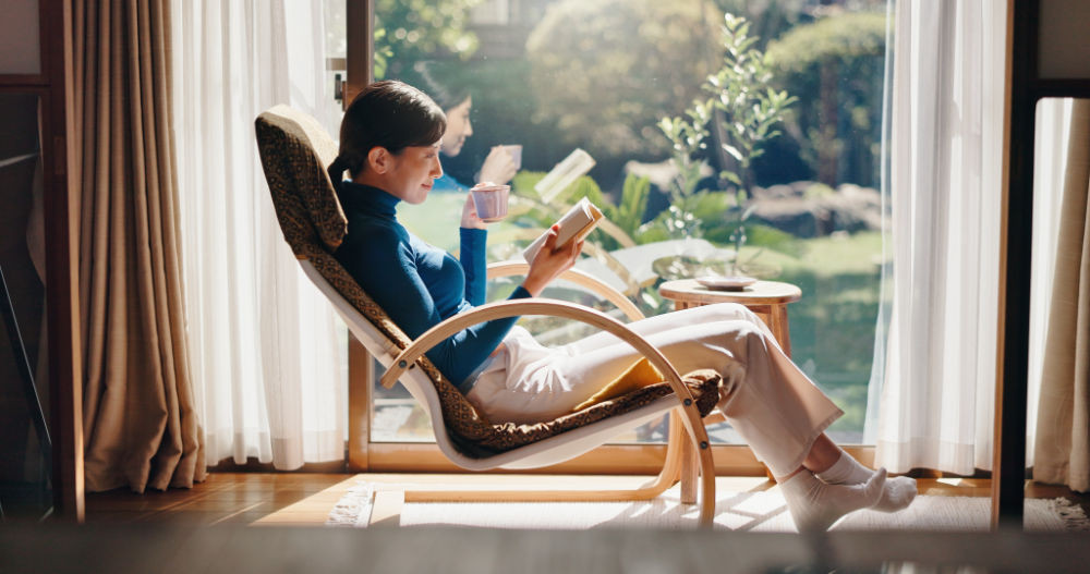A woman relaxing in a lounge chair by a sunlit window, holding a mug of coffee and a book.