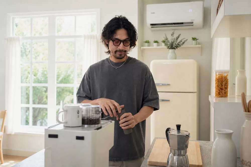 A man grinding coffee by hand in his kitchen, a moka pot and scale on the counter beside him.