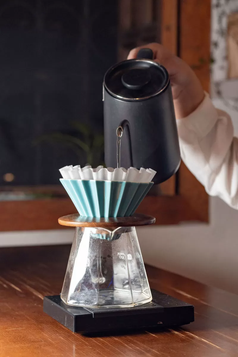 A hand pouring hot water from a kettle over a ceramic pour-over dripper set on a glass carafe